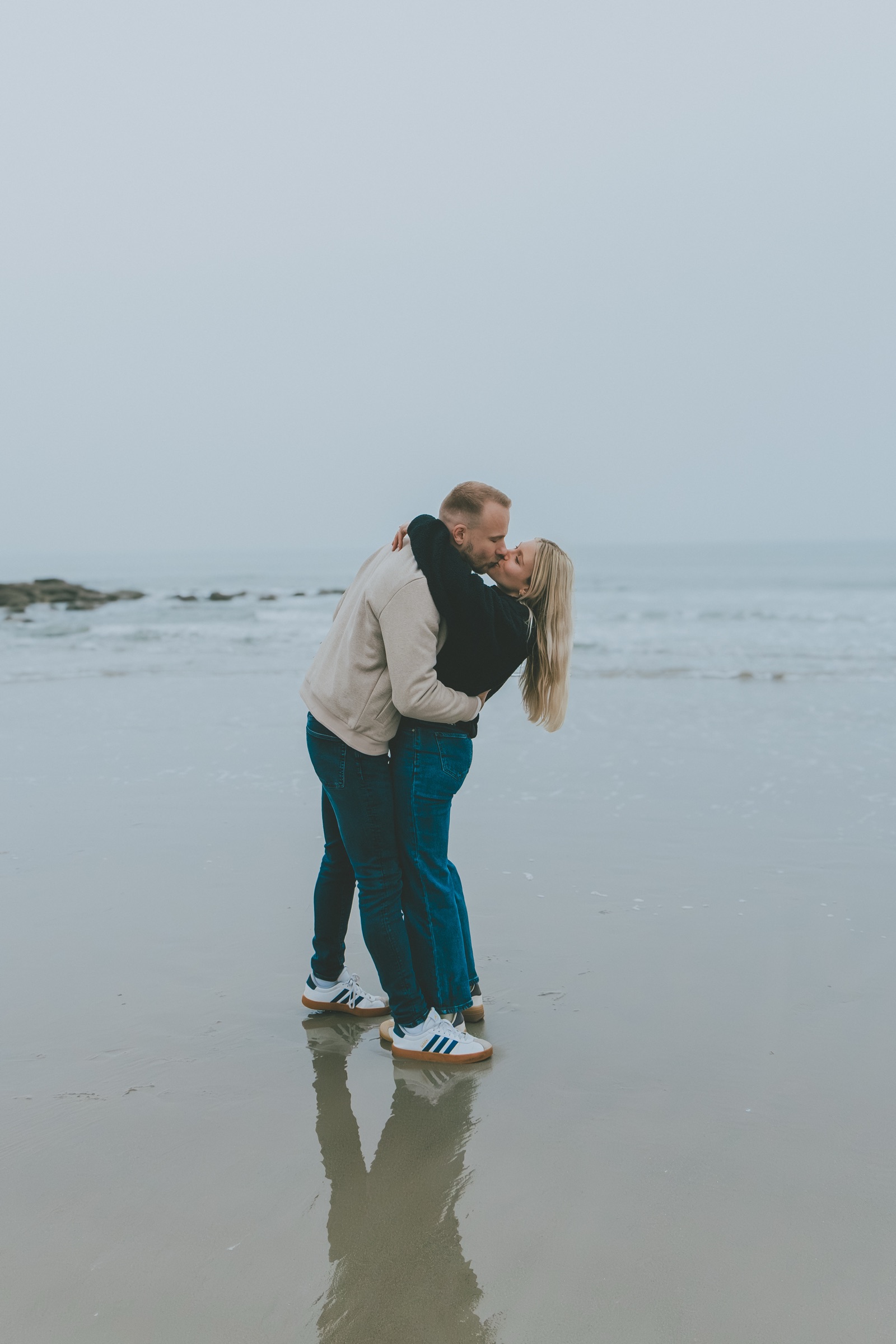 Amandine et Cyril s’embrassent sur la plage à marée basse, leur reflet sur le sable mouillé, l’horizon effacé par le brouillard