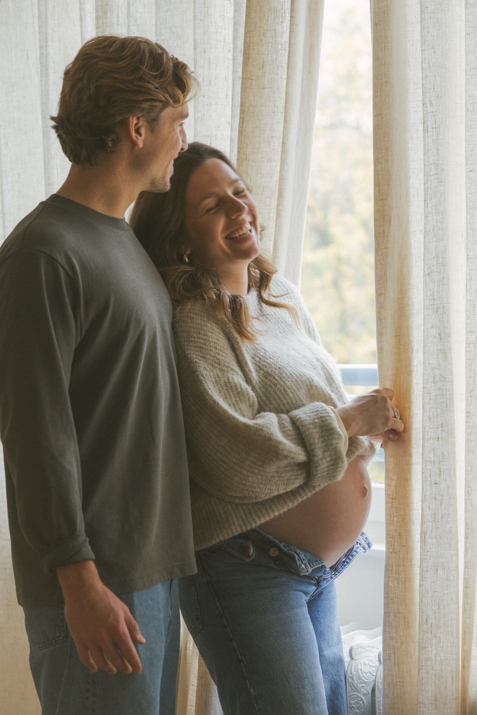 Futurs parents qui rient près d’une fenêtre lumineuse — séance photo de grossesse