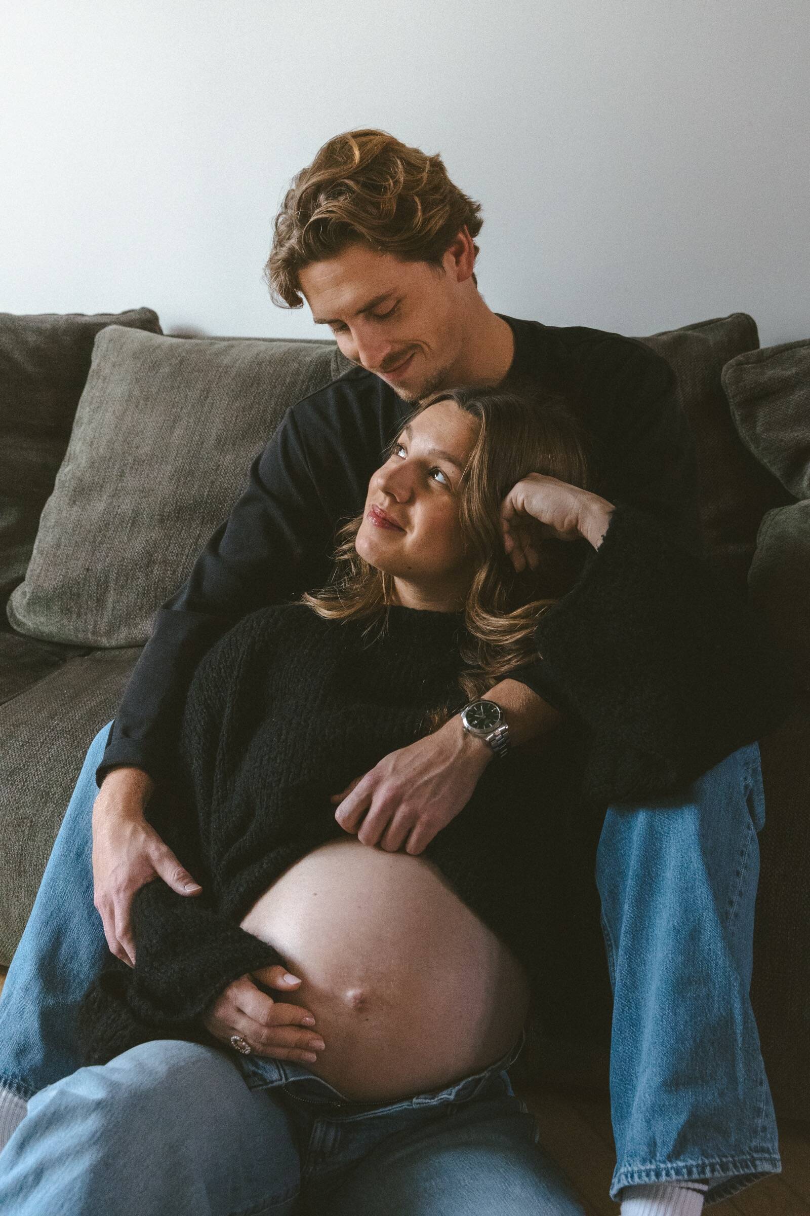 Futur papa enlaçant sa compagne enceinte sur un canapé, ventre arrondi — séance photo grossesse et naissance